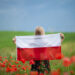 Blond girl holding flag of Poland in the poppy field. Back  view. Polish Flag Day. Independence Day. Travel and learn polish language concept.